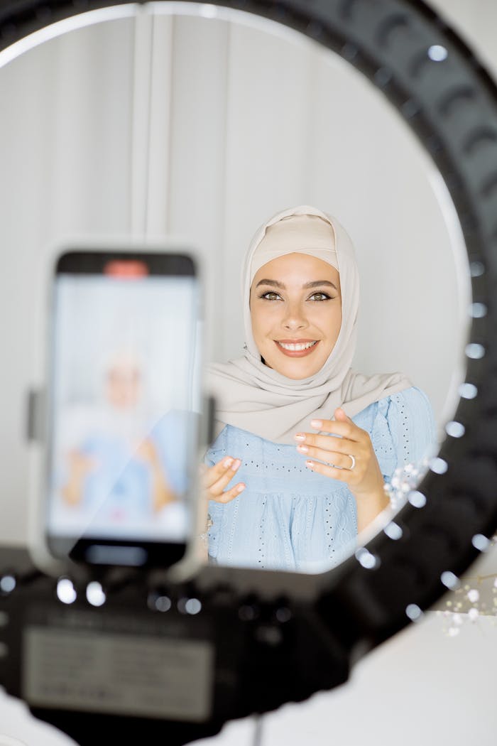 Smiling Muslim woman in hijab creating vlog content using a smartphone and ring light.